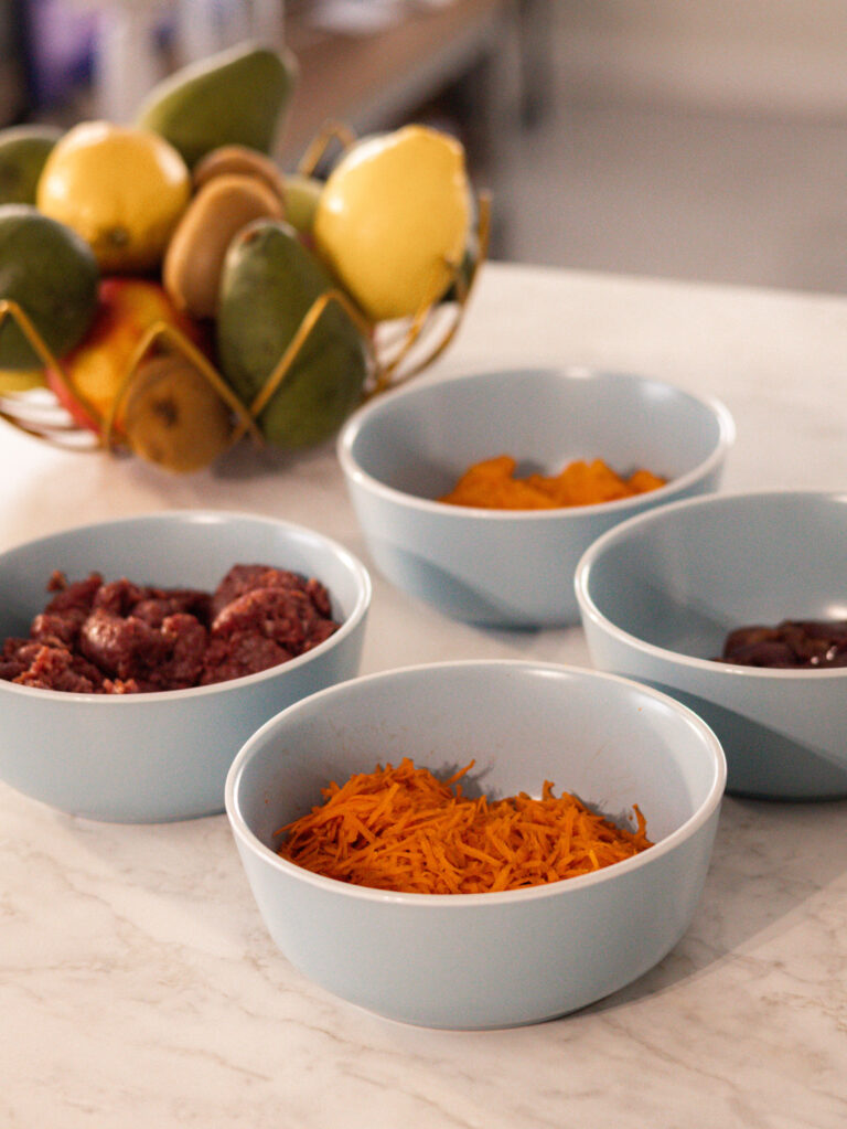 a table with bowls that have ingredients like chicken livers, butternut, carrots and raw meat
